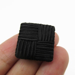 Könnte beinhalten: Ein schwarzer quadratischer Knopf aus gewebtem Stoff. Der Knopf hat ein strukturiertes, gewebtes Design.