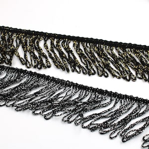 Peut inclure: Deux bandes de franges décoratives avec un motif en boucles. La bande supérieure est noire avec des fils dorés métallisés, tandis que la bande inférieure est noire avec des fils argentés métallisés. Les deux ont un bord tissé.