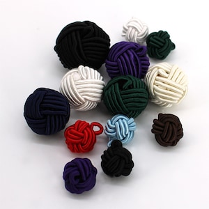 May include: A collection of spherical knot buttons in various sizes and colours, including black, white, navy blue, dark green, purple, red, light blue, and brown. Each button is constructed from tightly wound cord, featuring a textured surface.