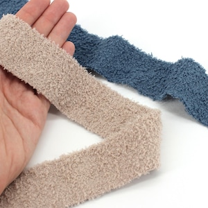 May include: Close-up of two fuzzy, textured headbands. One is a light beige color, and the other is a muted blue. The headbands are soft and plush, likely made of a cozy material.