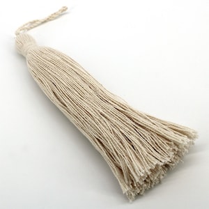 May include: A decorative tassel made of off-white cotton cord. The tassel features a tightly wrapped head section and a long, loose fringe of individual strands that fan out at the bottom. A small braided loop extends from the top for hanging.