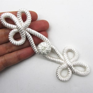 May include: White rope frog closure with a decorative knot and looped ends. This closure is a classic detail for coats, jackets, and other garments. The closure is made of a thick, woven rope.