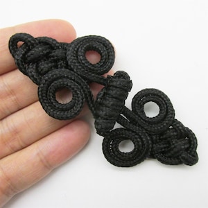 Puede incluir: Cierre de rana anudado negro, un cierre decorativo hecho de cordón tejido. El cierre presenta una serie de bucles y nudos, creando un diseño único e intrincado. El color es un negro profundo y sólido.