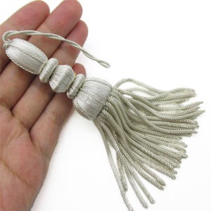 Puede incluir: Una borla decorativa de color plateado que presenta una cabeza de varias secciones con detalles acanalados y un fleco largo y fluido de hilos de cordón trenzado. La borla incluye un lazo de cordón plateado a juego en la parte superior para colgar.