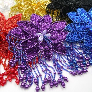 Può includere: Una collezione di applicazioni a forma di fiore con paillettes vivaci e frange di perline pendenti. Il set include pezzi singoli in viola, rosso, blu, oro, nero e bianco, ognuno con un centro di petali e fili di perline che pendono.
