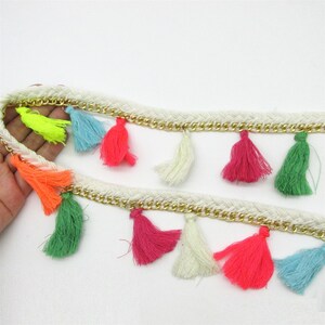 May include: A decorative trim featuring two parallel rows of braided white cord and gold-tone metal chain links. The trim is adorned with a series of small, dangling tassels in vibrant colours including neon yellow, orange, pink, green, and blue.