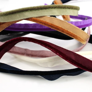 May include: A collection of velvet ribbons in various colors including olive green, tan, purple, deep red, cream, and navy blue. Each ribbon features a soft, textured velvet surface on one side and a smooth, flat finish on the reverse side.