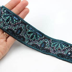 Può includere: Primo piano di un nastro decorativo con intricati abbellimenti di perline e paillettes. Il nastro presenta una base verde acqua scuro con motivi floreali in verde acqua e viola. Le paillettes e le perline aggiungono consistenza e brillantezza al design.