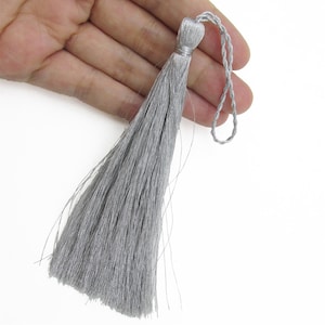 May include: A silver-coloured decorative tassel featuring a dense cluster of fine, shimmering threads hanging from a braided loop. The top of the tassel is tightly bound with matching silver thread, creating a smooth, cylindrical knot that transitions into long, straight, flowing strands.