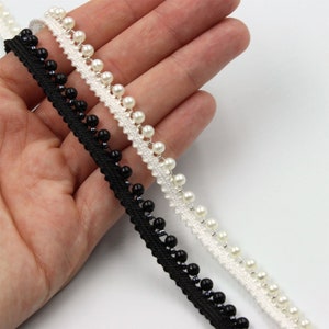May include: Two trims of black and white beaded lace with a scalloped edge. The lace is shown on a hand.