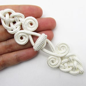 May include: A white cord frog closure fastener featuring intricate woven knot patterns and spiral designs. The fastener consists of two symmetrical pieces, each with a central knot and two coiled ends, connected by a central loop and toggle mechanism.