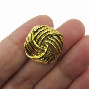 Peut inclure: Un bouton circulaire doré présentant une texture en relief en forme de spirale rappelant une corde qui rayonne depuis le centre. La surface métallique a une finition polie avec des rainures profondes entre les lignes courbes.