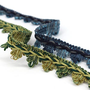 May include: Three different trims of braided and fringed fabric in shades of blue, green, and gold. The trims are woven with a rope-like texture and have a decorative fringe.