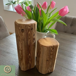 Puede incluir: Dos rebanadas de tronco de madera rústico, una más alta que la otra, con veta de madera natural y color marrón claro. Los troncos están dispuestos sobre una mesa de madera con un ramo de tulipanes rosas en un jarrón blanco.