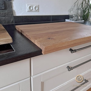 Può includere: Un grande tagliere in legno marrone chiaro si trova su un piano di lavoro nero. Il piano di lavoro fa parte di una cucina bianca con due cassetti e maniglie argentate. I cassetti hanno un design testurizzato in rilievo. Il tagliere ha un bordo leggermente arrotondato ed è fatto di legno naturale. Il nome del marchio "STEINHOLZ" è visibile nell'angolo in basso a destra dell'immagine.