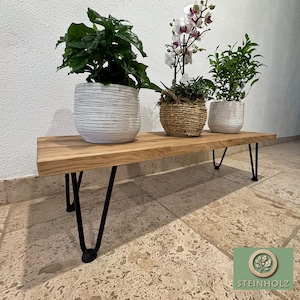 May include: A wooden bench with black hairpin legs, featuring three potted plants. The bench is made of natural wood with a light brown finish. The plants include a green leafy plant, a white orchid, and a green bush.