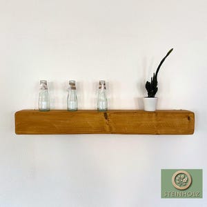 May include: A wooden shelf with a natural finish, featuring three clear glass bottles and a small potted plant. The shelf is mounted on a white wall.