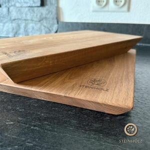 Può includere: Due taglieri in legno impilati uno sull'altro. La tavola superiore ha un bordo smussato. Le tavole sono in legno marrone chiaro e hanno la scritta "STEINHOLZ" e un logo inciso nel legno.