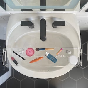 Puede incluir: Un lavabo de baño blanco con una bandeja de acrílico transparente que contiene varios productos de belleza, incluyendo un pincel de maquillaje, lápiz labial, bálsamo labial y una botella de protector solar. El texto "grateful heart" es visible en la bandeja.