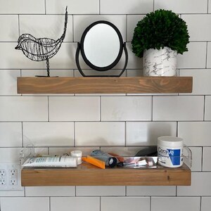 May include: Two horizontal wooden floating shelves mounted on a white tiled wall. The top shelf holds a black wire bird sculpture, a round black-framed mirror, and a green plant in a white marble-patterned pot. The bottom shelf is clear acrylic.