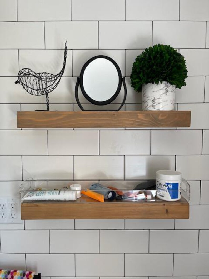 May include: Two horizontal wooden floating shelves mounted on a white tiled wall. The top shelf holds a black wire bird sculpture, a round black-framed mirror, and a green plant in a white marble-patterned pot. The bottom shelf is clear acrylic.