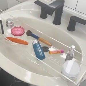 May include: A clear acrylic bathtub tray resting over a white pedestal sink with a matte black faucet. The tray holds various beauty products including a pink compact, an orange mascara tube, a makeup brush, a blue bottle, a lip balm, and jars.