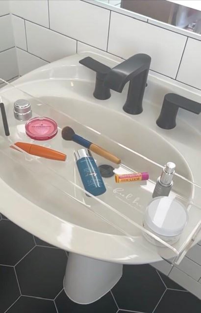 May include: A clear acrylic bathtub tray resting over a white pedestal sink with a matte black faucet. The tray holds various beauty products including a pink compact, an orange mascara tube, a makeup brush, a blue bottle, a lip balm, and jars.
