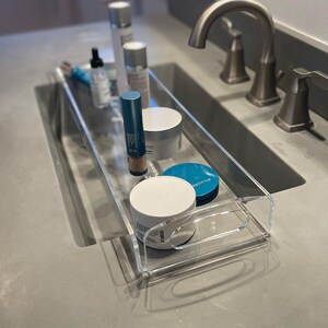 May include: A clear acrylic rectangular tray with integrated handles, containing various skincare products including cylindrical bottles, a dropper bottle, and round jars with white and teal lids, positioned on a gray countertop.