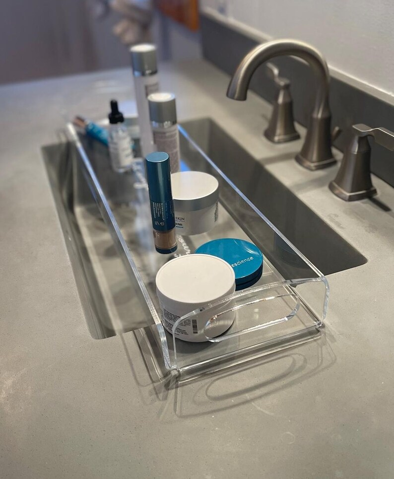 May include: A clear acrylic rectangular tray with integrated handles, containing various skincare products including cylindrical bottles, a dropper bottle, and round jars with white and teal lids, positioned on a gray countertop.