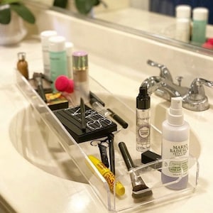 May include: A clear acrylic rectangular vanity tray holding various cosmetics including a black NARS palette, a Smashbox primer water spray, a Mario Badescu facial spray, a yellow mascara tube, makeup brushes, and other assorted beauty products.