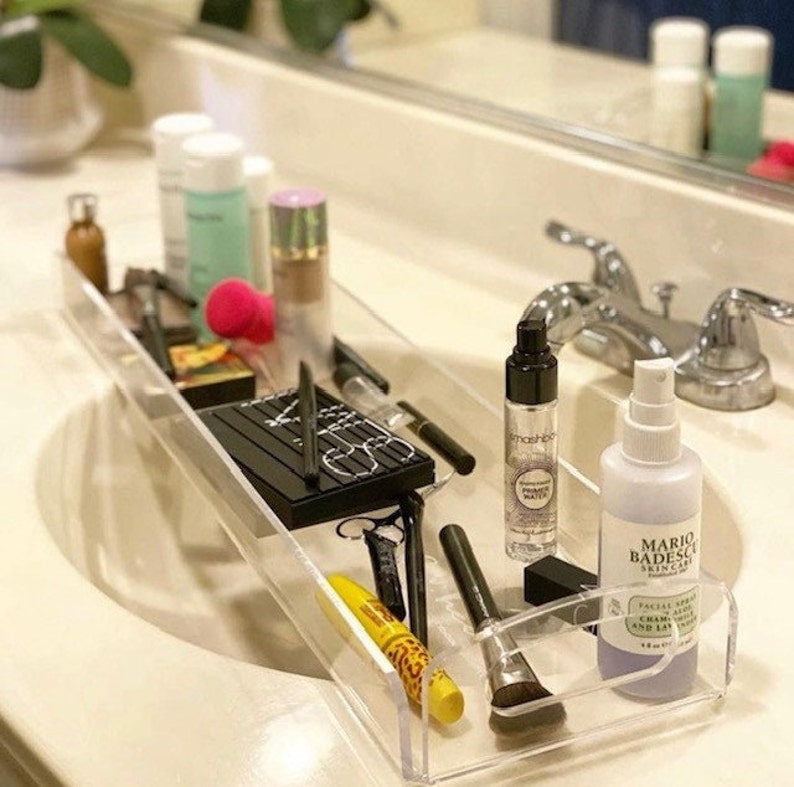 May include: A clear acrylic rectangular vanity tray holding various cosmetics including a black NARS palette, a Smashbox primer water spray, a Mario Badescu facial spray, a yellow mascara tube, makeup brushes, and other assorted beauty products.