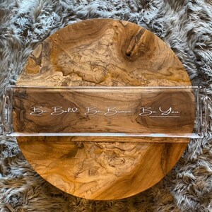 May include: A rectangular clear acrylic serving tray with integrated side handles. The tray features white cursive text printed on the surface that reads, Be Bold Be Brave Be You, positioned over a natural light brown wood grain circular base.