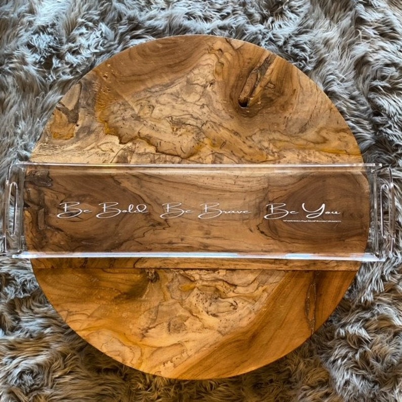 May include: A rectangular clear acrylic serving tray with integrated side handles. The tray features white cursive text printed on the surface that reads, Be Bold Be Brave Be You, positioned over a natural light brown wood grain circular base.