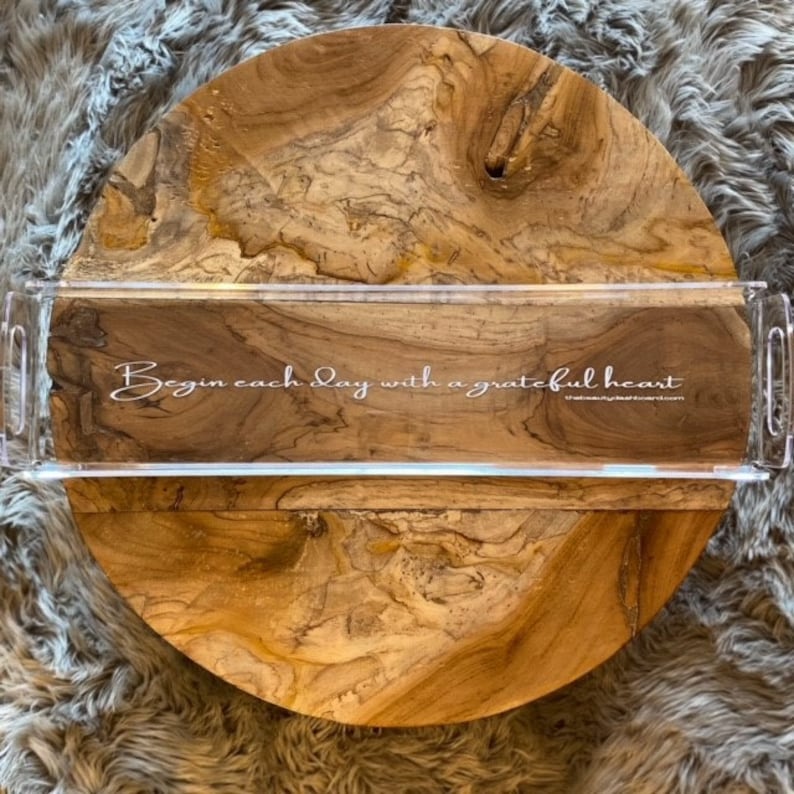 May include: A rectangular clear acrylic tray with side handles placed over a round wooden board with a natural grain pattern. The tray features the white printed text: Begin each day with a grateful heart, followed by thebeautyboard.com.