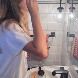May include: A clear acrylic bathroom vanity organizer tray containing various beauty tools, including a black hairbrush, a gold lipstick tube, a white jar, a yellow makeup brush, and a small hair clip, resting on a white porcelain sink basin.