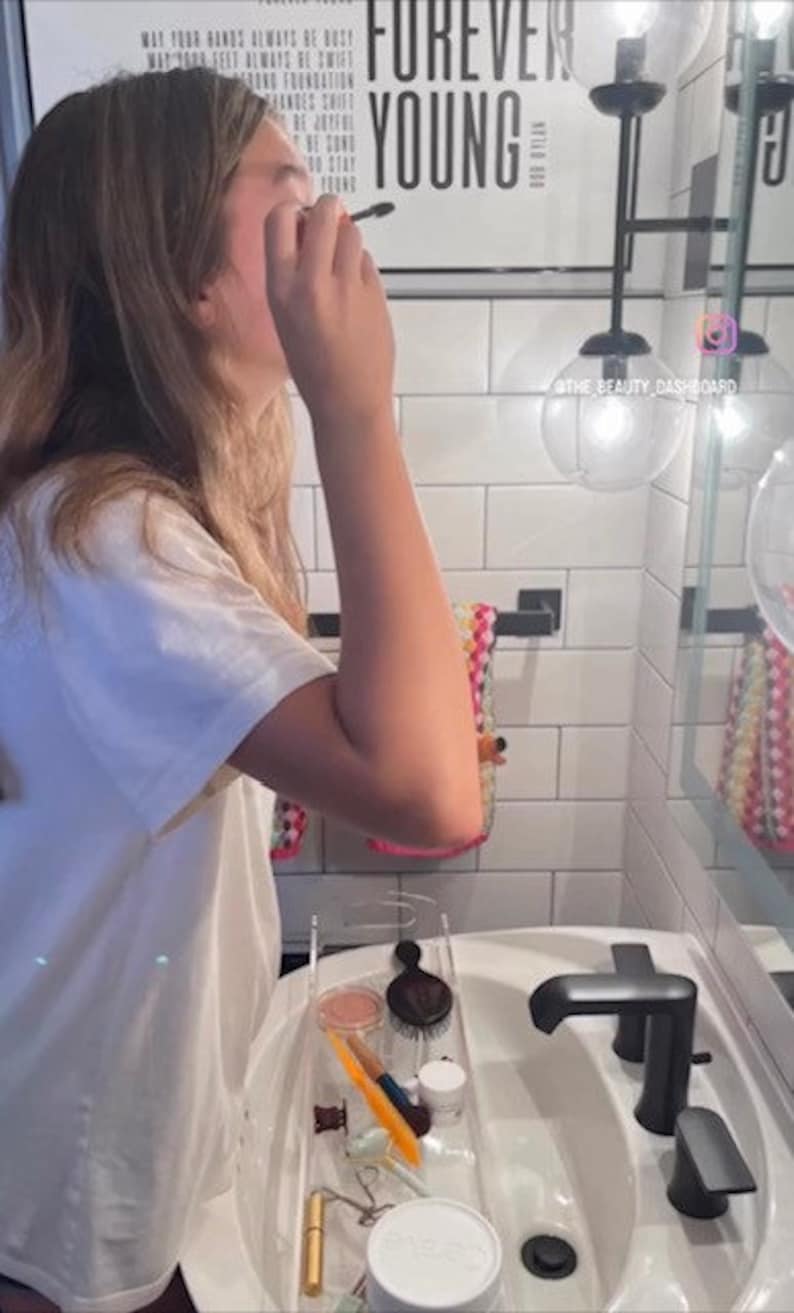 May include: A clear acrylic bathroom vanity organizer tray containing various beauty tools, including a black hairbrush, a gold lipstick tube, a white jar, a yellow makeup brush, and a small hair clip, resting on a white porcelain sink basin.