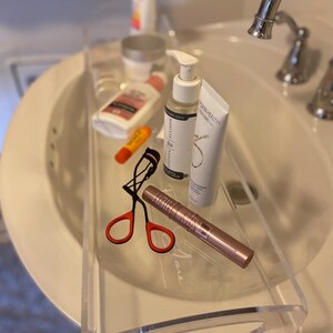 May include: A clear acrylic tray resting over a white bathroom sink, containing a black eyelash curler with orange handles, a rose gold mascara tube, a white pump bottle, a white tube of cleanser, a small orange lip balm, and a white lotion bottle.