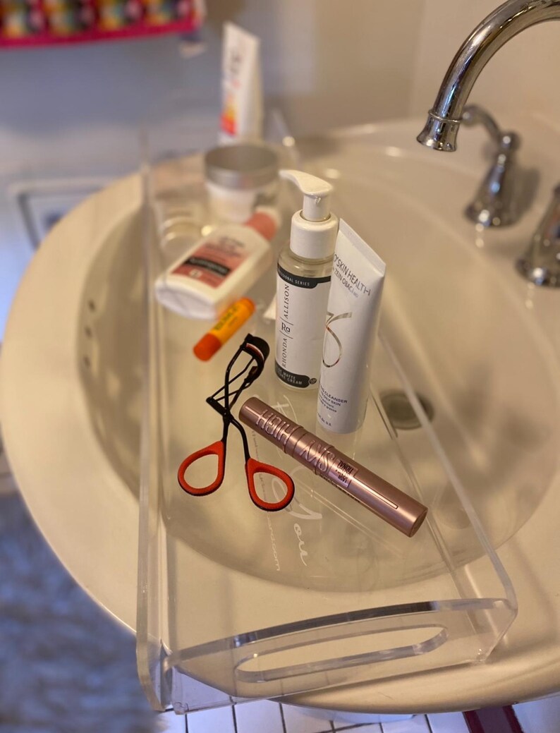 May include: A clear acrylic tray resting over a white bathroom sink, containing a black eyelash curler with orange handles, a rose gold mascara tube, a white pump bottle, a white tube of cleanser, a small orange lip balm, and a white lotion bottle.