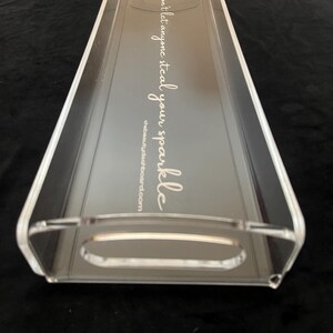 May include: A clear rectangular acrylic tray with integrated handles on both ends. The base features white cursive text that reads, "Don't let anyone steal your sparkle," followed by the website address, thebeautydashboard.com, printed in a smaller font.