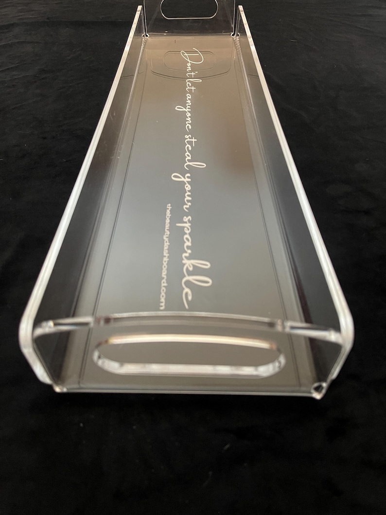 May include: A clear rectangular acrylic tray with integrated handles on both ends. The base features white cursive text that reads, "Don't let anyone steal your sparkle," followed by the website address, thebeautydashboard.com, printed in a smaller font.