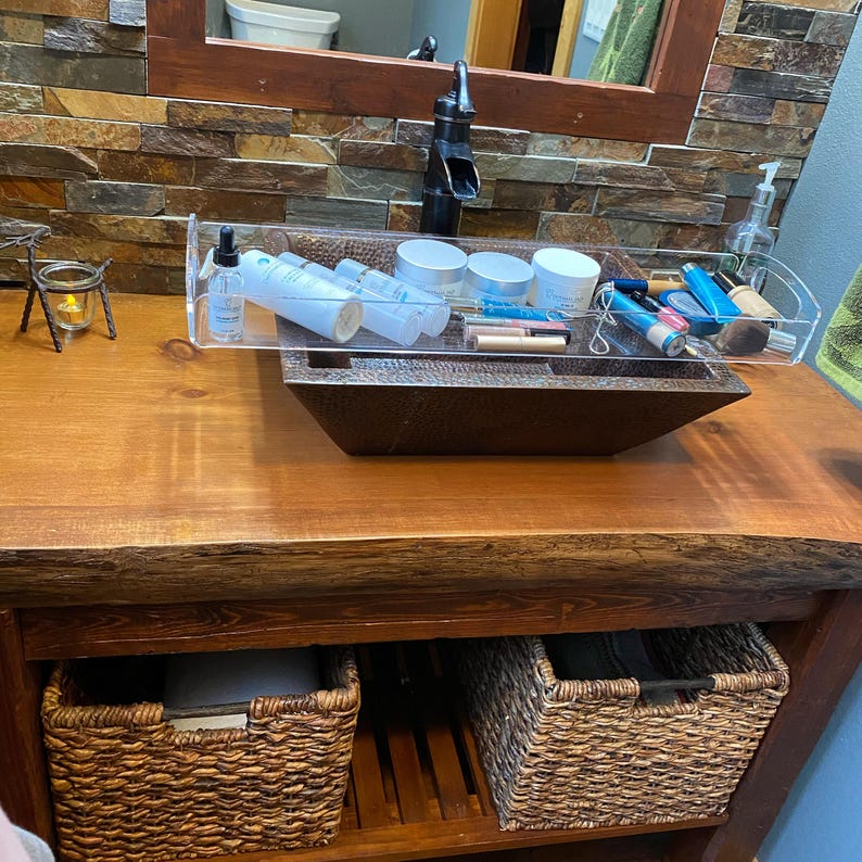 May include: A clear acrylic rectangular vanity tray holding various skincare and cosmetic products, including white jars, bottles, and makeup brushes, positioned over a dark brown hammered metal sink basin on a wooden bathroom vanity counter.