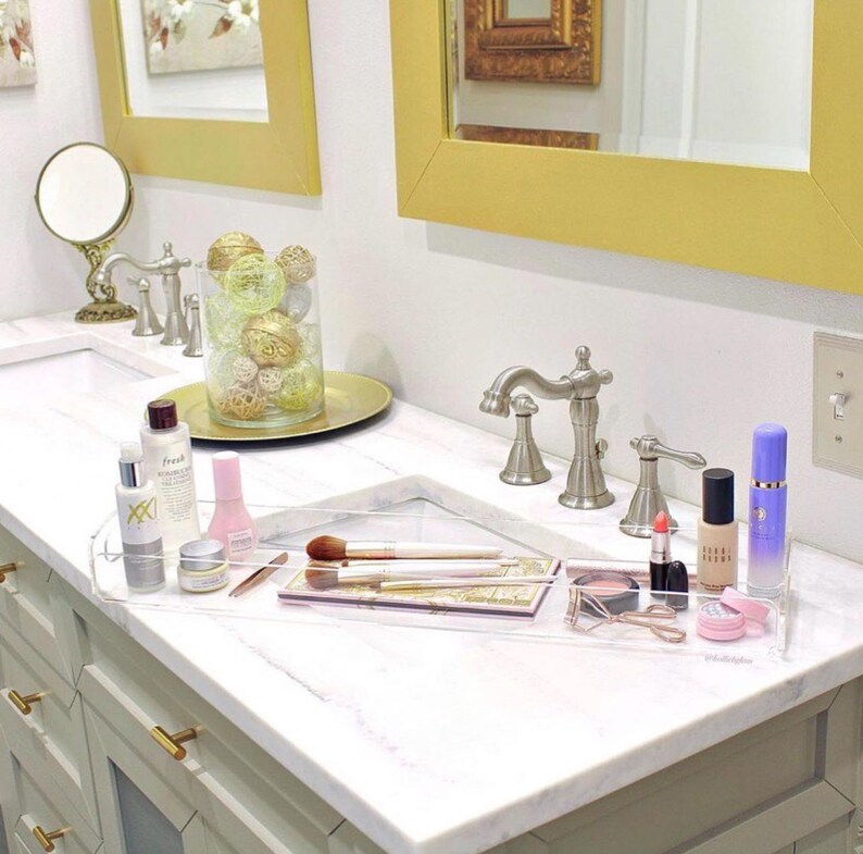 May include: A clear acrylic rectangular vanity tray holding various makeup items including brushes, lipsticks, a compact, and a lash curler. The tray sits on a white marble countertop next to a glass vase filled with decorative gold and silver spheres.