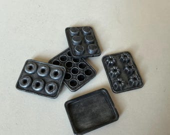 May include: A collection of miniature metal baking pans in various shapes, including donut, muffin, and cake molds, and a rectangular tray. The pans are a dark, weathered gray color, suggesting a vintage or rustic aesthetic.
