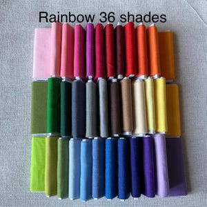 Puede incluir: Una colección de 36 carretes de hilo y cuadrados de fieltro a juego en un arcoíris de colores. El texto "Rainbow 36 shades" se muestra en la parte superior. Los colores van del rosa al morado, con tonos de rojo, naranja, amarillo, verde, azul y marrón.