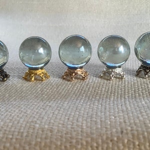 Puede incluir: Cinco orbes de vidrio sobre soportes decorativos. Los orbes son transparentes y tienen un ligero brillo iridiscente. Los soportes son de metal y están acabados en oro, plata y negro.