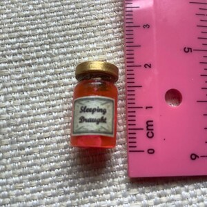 May include: A small glass jar with a gold lid filled with a red liquid. The jar has a label that reads "Sleeping Draught".