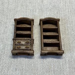 May include: Two miniature wooden bookshelves. One has two drawers with gold-colored knobs. The other is a simple bookshelf with shelves. Both are brown and have a classic design, perfect for a dollhouse or miniature collection.