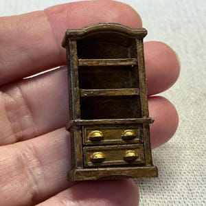 May include: A miniature wooden bookcase with a brown finish and gold-colored drawer pulls. The bookcase has three shelves and two drawers.