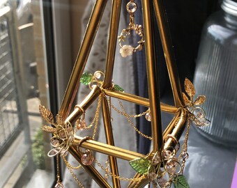 May include: A gold geometric mobile with delicate floral accents and crystal beads hanging from chains. The mobile is made from gold-painted drinking straws and features small green leaves and pink flowers.