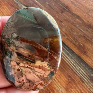 May include: A polished oval-shaped stone with a mix of brown, red, and green colors. The stone has a swirling pattern and a smooth, glossy finish.
